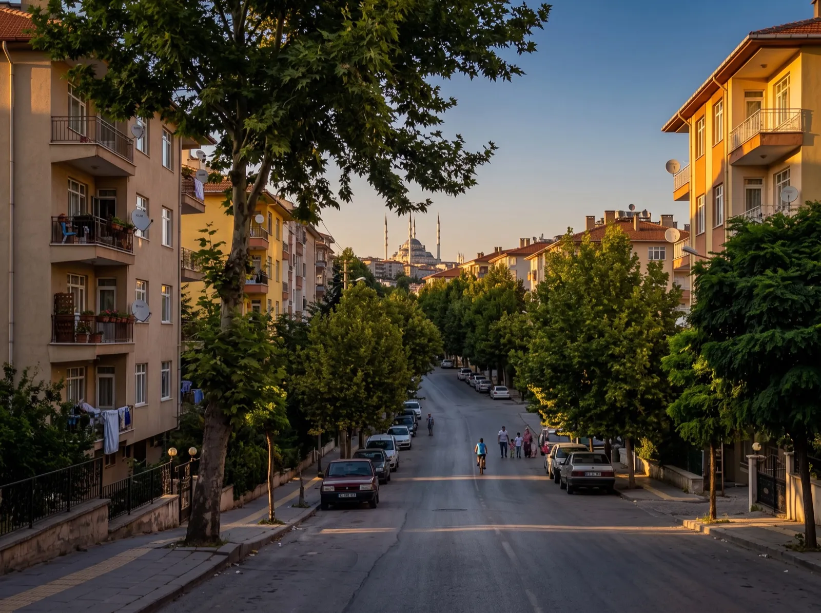 İki yanında apartmanlar ve ağaçlar bulunan, akşamüstü yürüyen insanların olduğu sakin bir mahalle sokağı